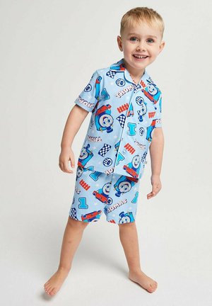 Smiling young boy in blue short-sleeve shirt and shorts set covered in colorful Thomas the Tank Engine prints stands barefoot on plain background.