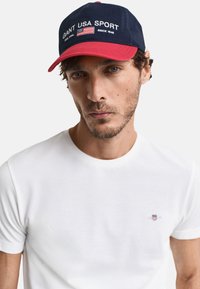 Marineblå og rød baseballcap med broderet "GANT USA SPORT" og amerikansk flag. Båret med en simpel hvid t-shirt med korte ærmer i bomuldsstof.