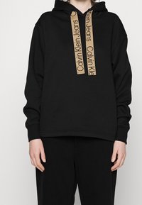 Czarna bluza z kapturem o luźnym kroju, z beżowymi sznurkami z logo i ściągaczami przy rękawach. Gładka tkanina o minimalistycznym designie.