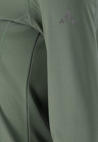 Grön långärmad plagg gjort av ett smidigt, elastiskt tyg. Har en diskret logotyp i grått på ärmen och flatlock-sömmar.