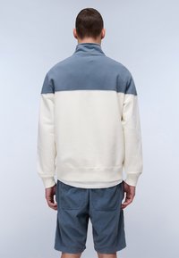 Light-colored sweatshirt with a blue upper section, high collar, and ribbed cuffs. Paired with blue shorts, textured fabric, relaxed fit.