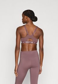 Καφέ αθλητικός σουτιέν με ρυθμιζόμενες τιράντες και σχέδιο με άνοιγμα, με λευκή κορδέλα με το λογότυπο. Συνδυάζεται με κολάν ψηλόμεσο στο ίδιο χρώμα.