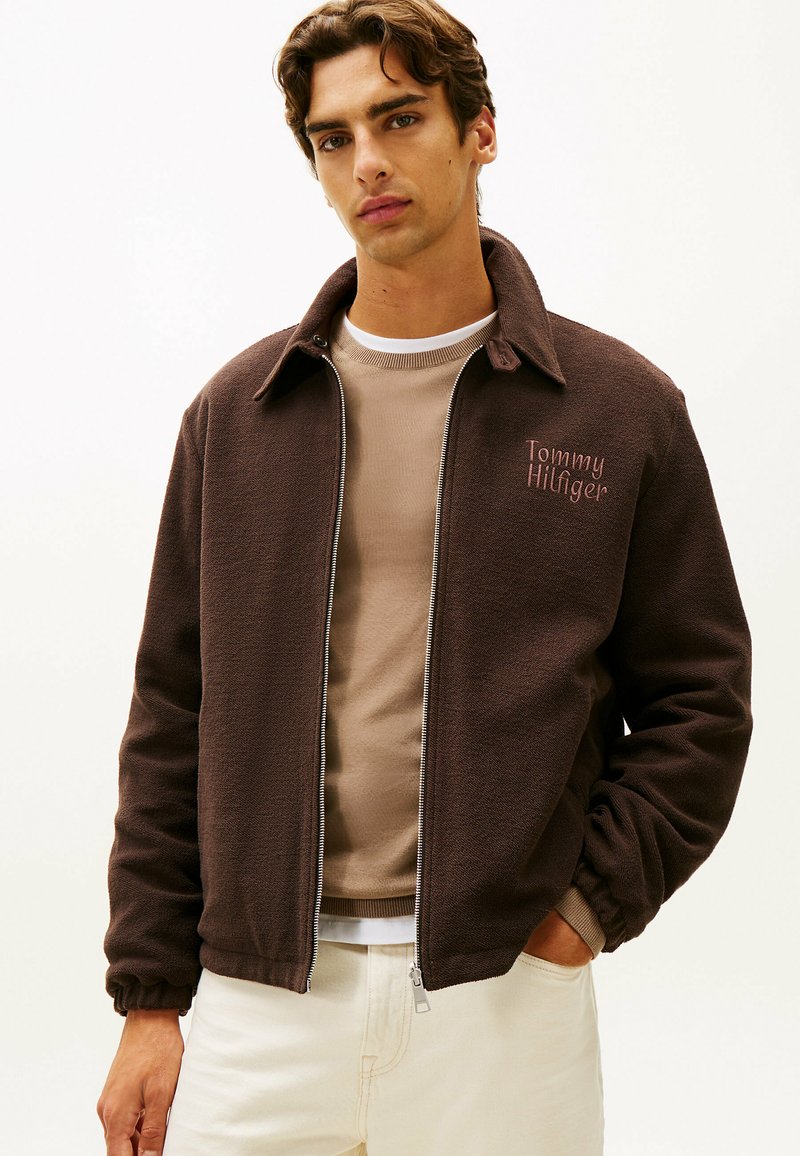 Brązowa polarowa kurtka na zamek z kołnierzykiem, z różowym haftem logo Tommy Hilfiger na lewym piersi, noszona na beżowej koszuli.