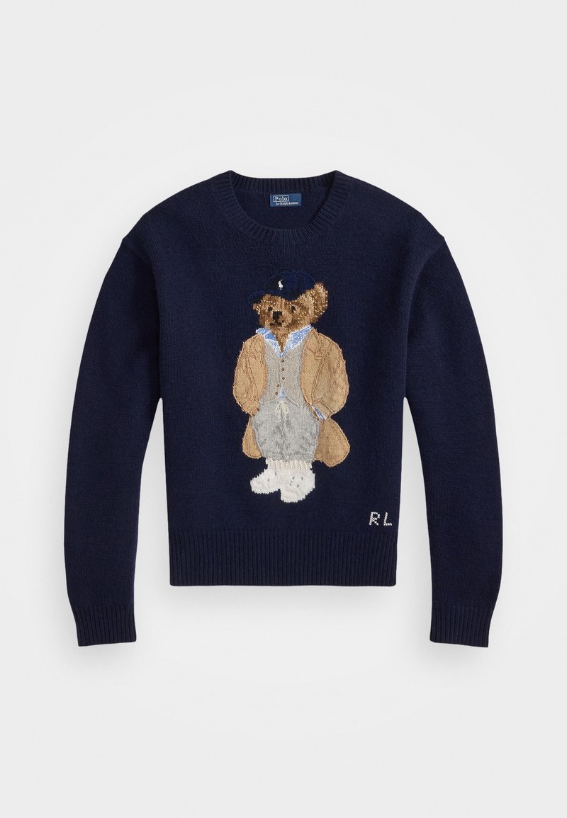 Maglione navy con un orso ricamato in dettaglio, che indossa un cappotto beige e pantaloni grigi, con polsini a coste e scollatura a girocollo.