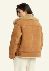 Bruin schapenvachtjack met een fluffy beige kraag, suède textuur, leren accenten en een zijgesp detail, gezien vanuit de achterkant.