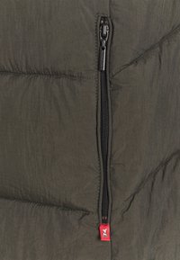Sección de chaqueta acolchada verde oscuro con una textura suave, que presenta un destacado zipper negro y una pequeña etiqueta de marca roja.