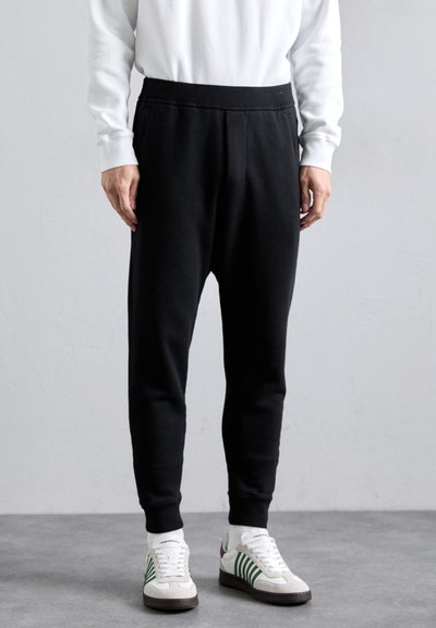 Pantaloni della tuta neri realizzati in un materiale morbido, con un design affusolato, cintura elastica e cuciture laterali. Indossati con scarpe da ginnastica bianche e verdi.