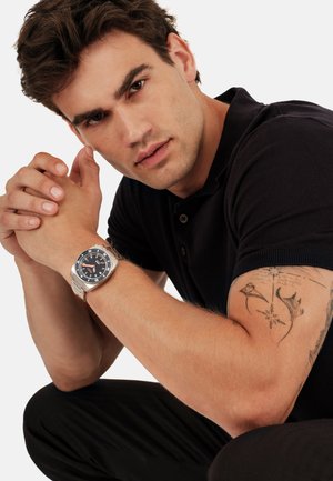 Ung mand iført sort polo-shirt med tatoveringer på venstre arm, bærende på et sølvfarvet armbåndsur, læner sig frem med hænderne foldet.