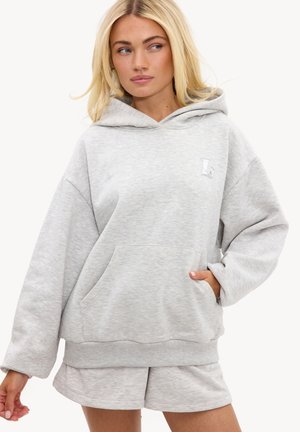 Femme aux cheveux blonds portant un sweat à capuche oversize gris clair et un short assorti, regardant sur le côté avec une main dans la poche du sweat.