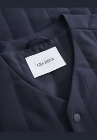 Navy blue quilted fabric with a prominent collar. Features a white label with "LES DEUX" printed in black, and a round black button accent.