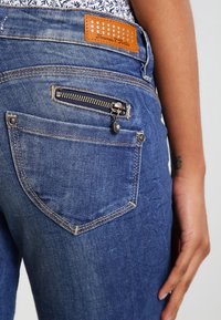 Jeans i denim har en mörkblå tvätt med en bakfickes dragkedja och en brun läderlapp. Synlig sömnad och detaljer i hårdvara.