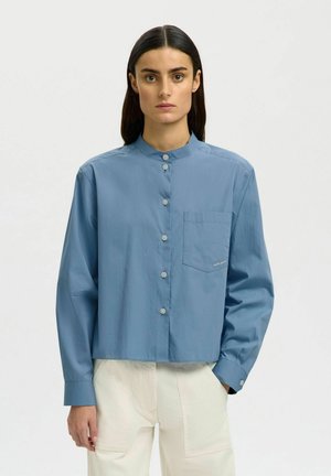 Kvinde med langt mørkt hår iført en blå skjorte med knapper og mandarinkrave samt cremefarvede bukser med høj talje, stående foran en ensfarvet baggrund.