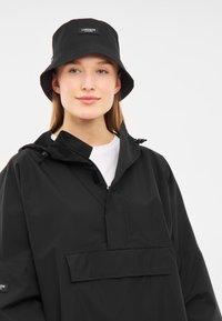 Musta vettä hylkivä ämpärihattu, jossa on logopatchi, yhdistettynä mustaan pullover-takkiin, jossa on etutasku ja korkea kaulus.