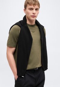 Νεαρός άνδρας με ελαφρύ πουλόβερ σε χακί χρώμα με κοντά μανίκια και λογότυπο «BOSS», μαύρο παντελόνι και μαύρο πουλόβερ περασμένο στους ώμους.