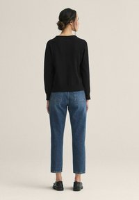 Nainen, jolla on tummat hiukset nutturalla, musta neulepaita, siniset farkut ja mustat kengät, seisoo selkä kameraan päin yksinkertaista taustaa vasten.