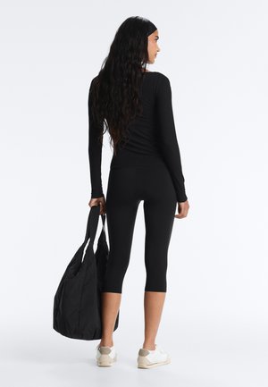 Kobieta o długich czarnych włosach, ubrana w czarną, długorękawową bluzkę i krótkie legginsy, trzymająca dużą czarną torbę na ramię, ubrana w białe trampki, stojąca bokiem.