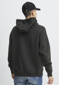 Felpa con cappuccio grigio scuro, dotata di un ampio cappuccio, polsini e orlo a coste. Indossata con jeans blu. La vista mostra la parte posteriore del capo e del cappello.