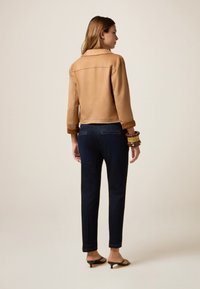 Giacca in suede marrone chiaro con colletto e maniche corte, abbinata a jeans tapered blu navy. Gli accessori includono bracciali impilati e sandali.