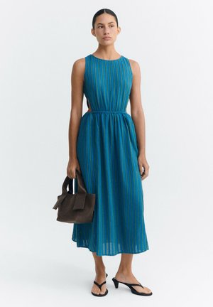 Frau mit dunklen Haaren in einem blau gestreiften ärmellosen Midikleid, die eine braune Handtasche hält und schwarze offene Sandalen mit Absatz trägt, stehend vor weißem Hintergrund.