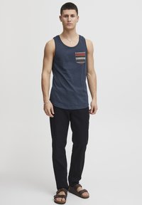 Maiou bleumarin cu o buzunar cu model, pantaloni negri și sandale maro. Materialul pare moale; maioul are un decolteu rotunjit și design fără mâneci.