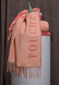 Couverture tissée pêche avec bords frangés, présentant "TOUCHÉ" en lettres roses audacieuses, drapée sur un présentoir cylindrique gris uni.