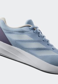 Hellblaues Sportschuhmodell mit einem Obermaterial aus Mesh, strukturierten Einsätzen, weißer Sohle und lila Akzent an der Ferse. Verfügt über passende Schnürsenkel und Logo.