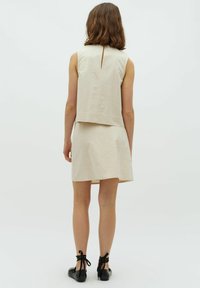 Beige, ärmelloses Oberteil mit Schlitz am Rücken, kombiniert mit einem passenden Rock. Glatte Textur, minimalistisches Design und schlichte Silhouette. Schwarze Schnürschuhe.