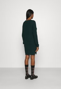 Vestido de malha em verde escuro com gola redonda, mangas longas e fendas laterais. Combinado com meias de xadrez até ao joelho e sapatos pretos.