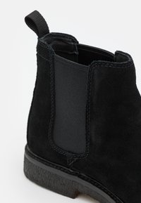Schwarze Wildleder Chelsea-Boots mit elastischem Seiteneinsatz, Zugschlaufe auf der Rückseite, detaillierter Nahtverarbeitung und strukturiertem Gummisohle auf weißem Hintergrund.