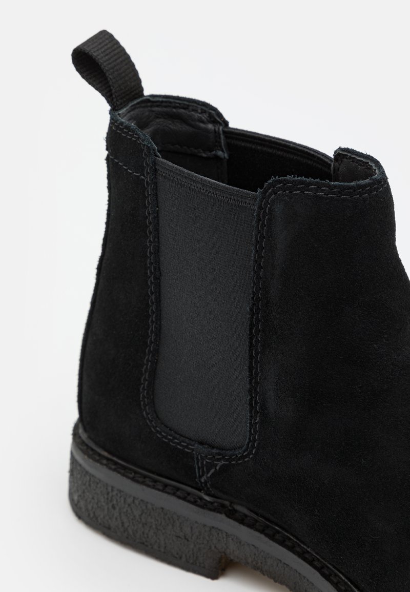 Bottines Chelsea en daim noir avec panneau élastique sur le côté, languette à l'arrière, couture détaillée et semelle en caoutchouc texturée sur fond blanc.