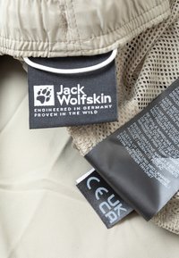 Lichtbeige stof met gaasdetails. Labels bevatten het "Jack Wolfskin"-logo, verzorgingsinstructies en certificeringsdetails in meerdere talen.