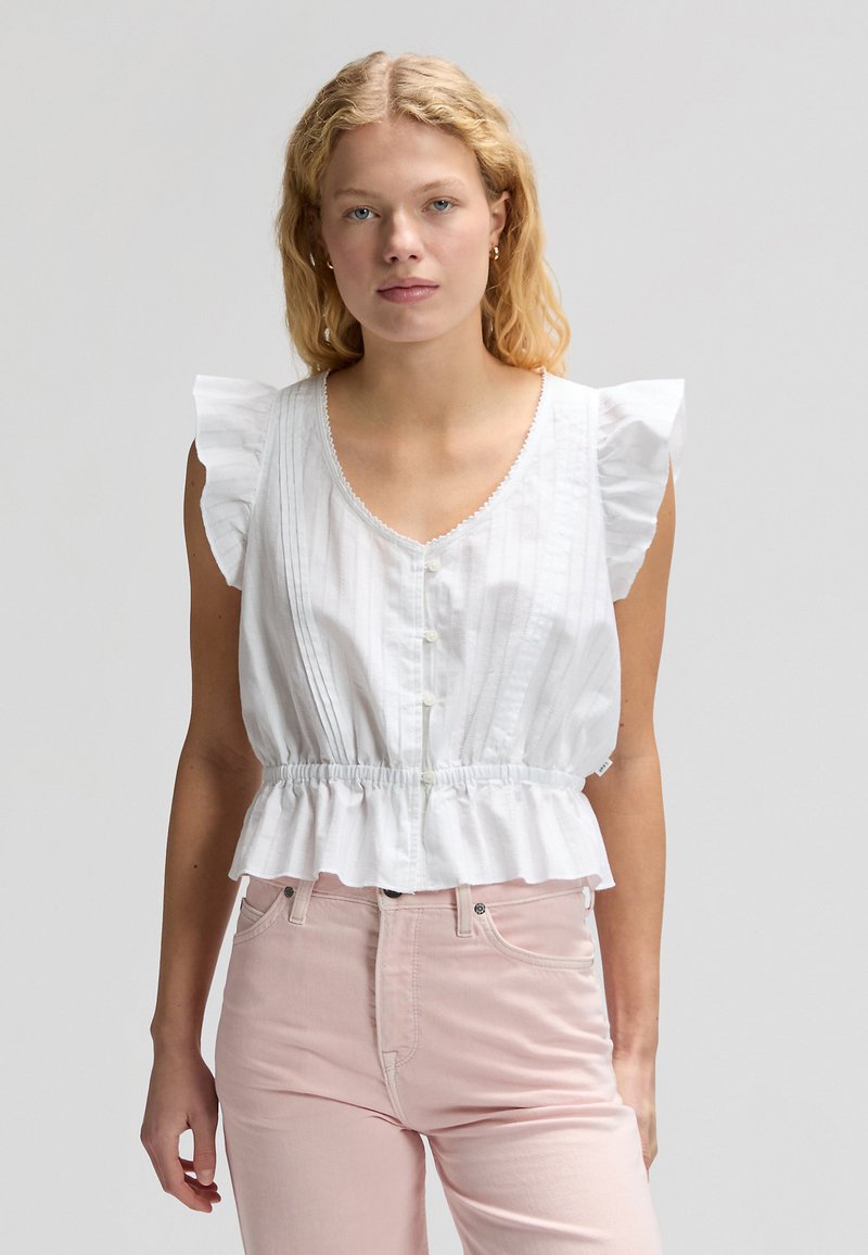 Νεαρή γυναίκα με ξανθά μαλλιά που φοράει λευκή αμάνικη μπλούζα με βολάν και ανοιχτό ροζ ψηλόμεσο παντελόνι, πάνω σε απλό φόντο.