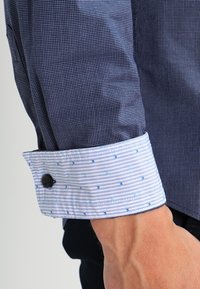 Rękaw niebieskiej koszuli w krateczkę z kontrastowym białym mankietem, na którym znajdują się małe niebieskie wzory i czarny guzik, spoczywający w pobliżu dłoni.