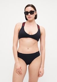 Svart bikini-sett med teksturert stoff, med brede stropper med rød "HUGO"-merkevare og et enkelt, lavt Design på bunnen. Svart solbriller.