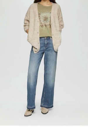 Persoon draagt een olijfgroen grafisch T-shirt, een beige gebreid vest met knopen, blauwe wijdvallende jeans en beige enkellaarzen, staand tegen een effen achtergrond.