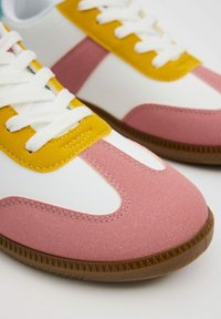 Zapatillas con una base blanca, acentos en rosa y amarillo. Punta de goma texturizada en rosa, cordones blancos y suela de goma marrón con un patrón de zigzag.