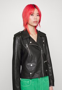 Svart läderbikerjacka med silverfärgad dragkedja och knappar, med två sidofickor och en kort design. Modell som bär gröna byxor.