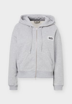 Svetlo siva jopa z zadrgo, z vrvico za zategovanje, žepi spredaj, rebrastimi manšetami in spodnjim robom ter majhno belo logotipno značko na levi strani prsnega koša.