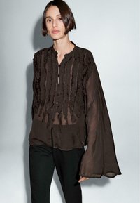 Donna dai capelli corti e scuri, con una blusa nera trasparente con ruffles verticali e maniche lunghe a campana, abbinata a pantaloni neri.