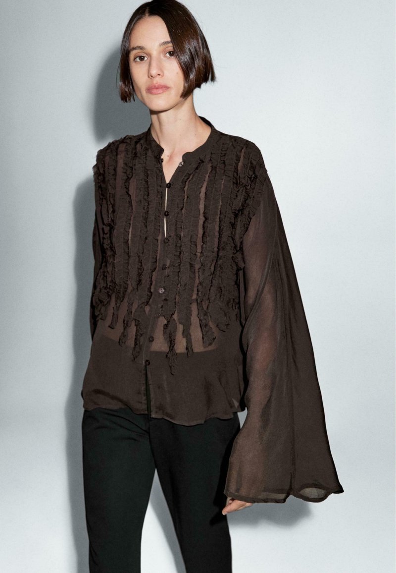 Donna dai capelli corti e scuri, con una blusa nera trasparente con ruffles verticali e maniche lunghe a campana, abbinata a pantaloni neri.