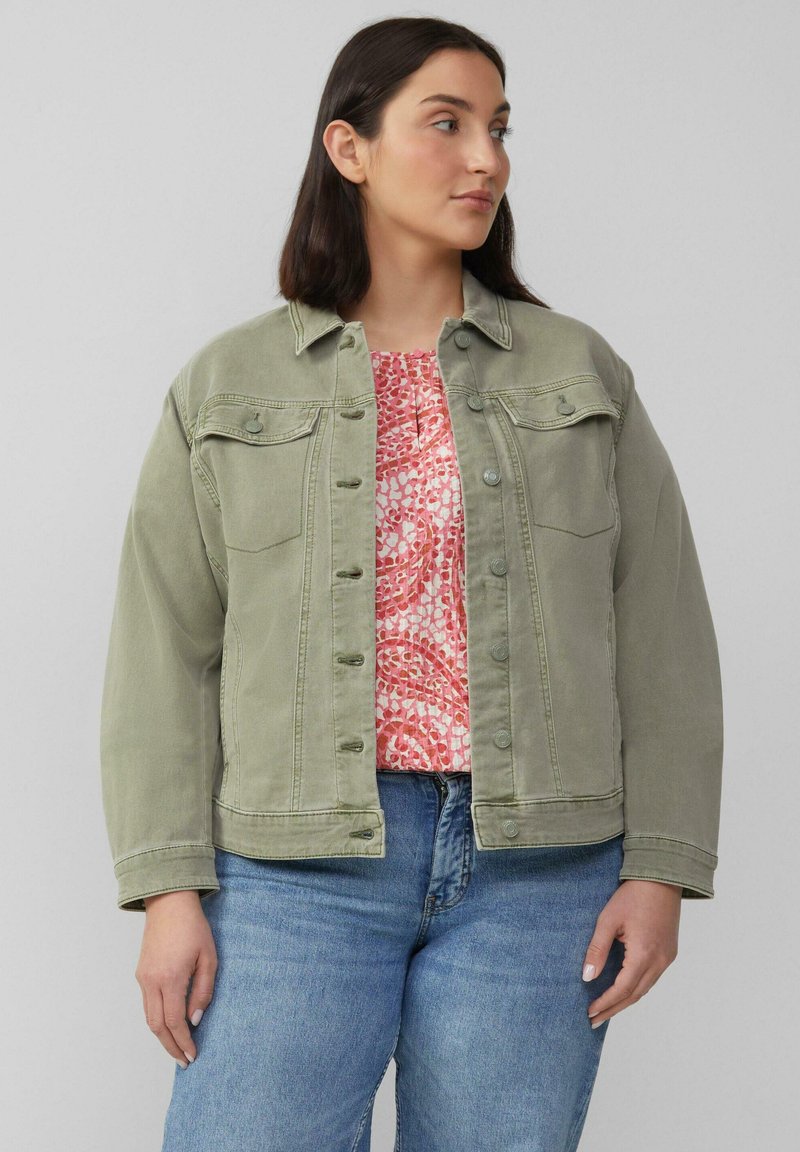 Olivengrüne Jeansjacke mit Knopfleiste, Brusttaschen und leicht lockerer Passform, die über einer rosa gemusterten Bluse getragen wird.