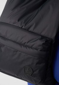 Bolso acolchado negro con una textura suave, que presenta un bolsillo frontal y un logo circular negro. Forma estructurada con sutiles detalles de costura.