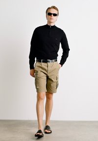 Tricou lung cu fermoar negru, pantaloni scurți cargo bej cu buzunare laterale, curea neagră, ochelari de soare și sandale negre, pe o podea gri împotriva unui perete alb.