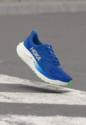 Kék HOKA futócipő fehér és zöld talppal, a levegőben aszfaltúton fehér csíkozással, kis törmelékeket felkavarva.