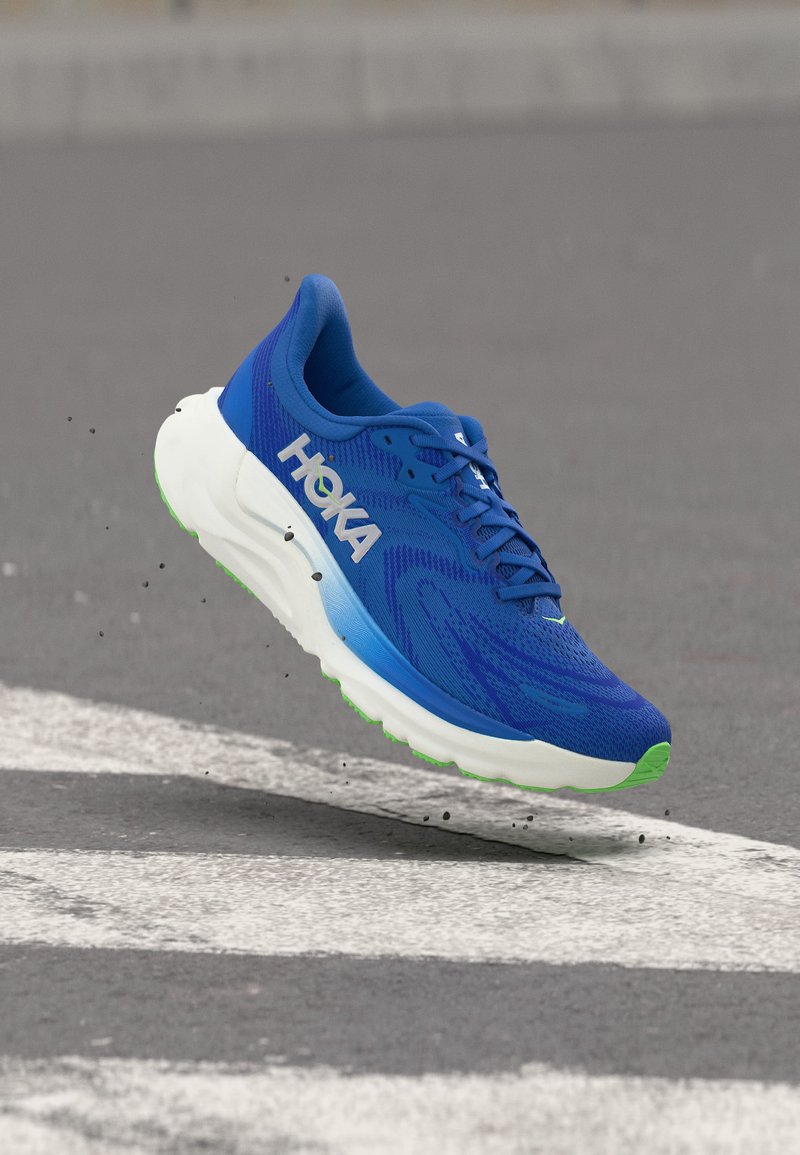 Μπλε αθλητικό παπούτσι HOKA με λευκή και πράσινη σόλα, στον αέρα πάνω από έναν ασφαλτοστρωμένο δρόμο με άσπρες λωρίδες, σηκώνοντας μικρά συντρίμμια.