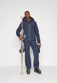 Casaco de esqui azul-marinho com fecho à frente e capuz, combinado com calças a condizer. Esquis e fixações na mão, a usar botas de esqui pretas.