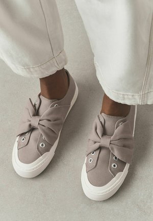 Fødder iført beige slip-on lærredssko med store stofsløjfer, sat sammen med lyse vidde bukser på en grå gulv.