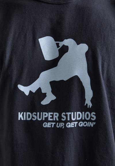 Sort bomulds T-shirt med et stort gråt grafik af en figur, der holder en krukke, med teksten "KIDSUPER STUDIOS" og "GET UP, GET GOIN'" nedenfor.