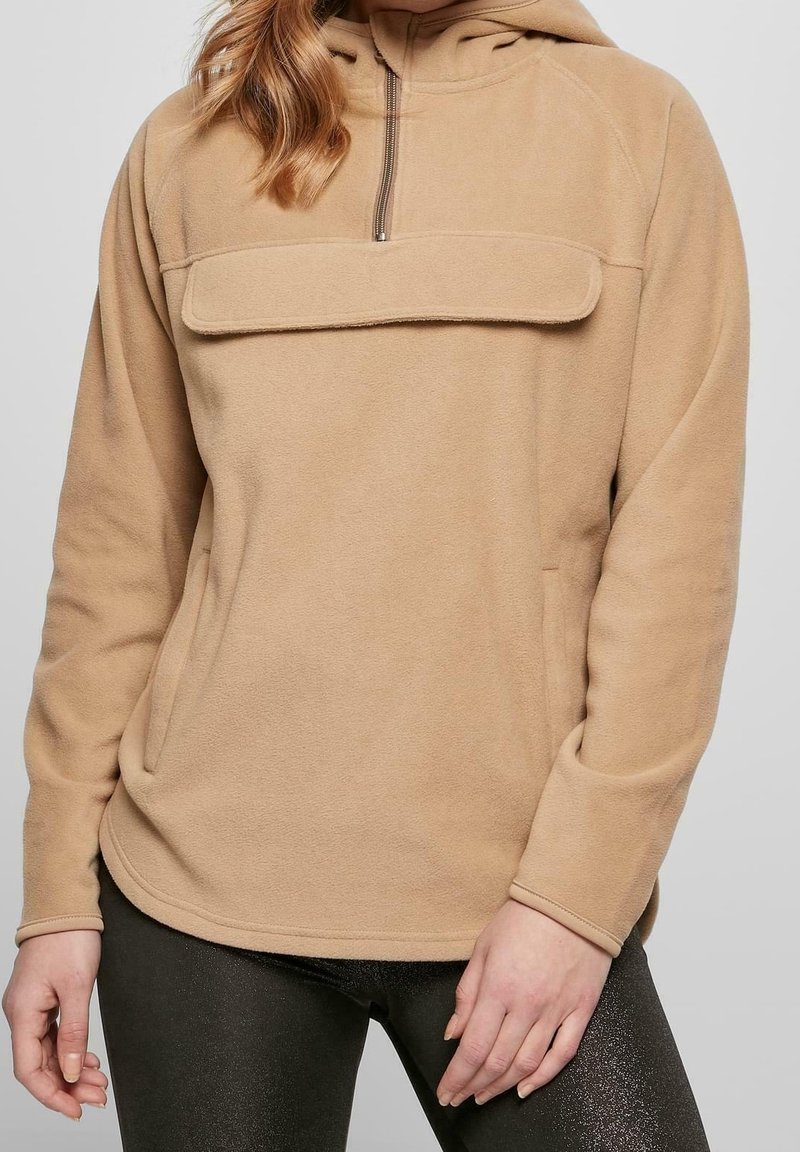 Femme portant un pull polaire beige avec zip devant et grande poche, associé à un legging noir sur un fond uni.