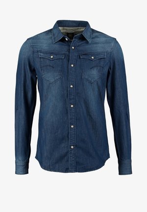Manteau long noir sur une chemise en denim bleu, associé à un jean noir et des chaussures foncées. Les caractéristiques incluent un col, une fermeture éclair sur le devant et des poches.